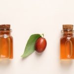 Diferentes botes de aceite de jojoba con hojas y semillas sobre fondo blanco roto.