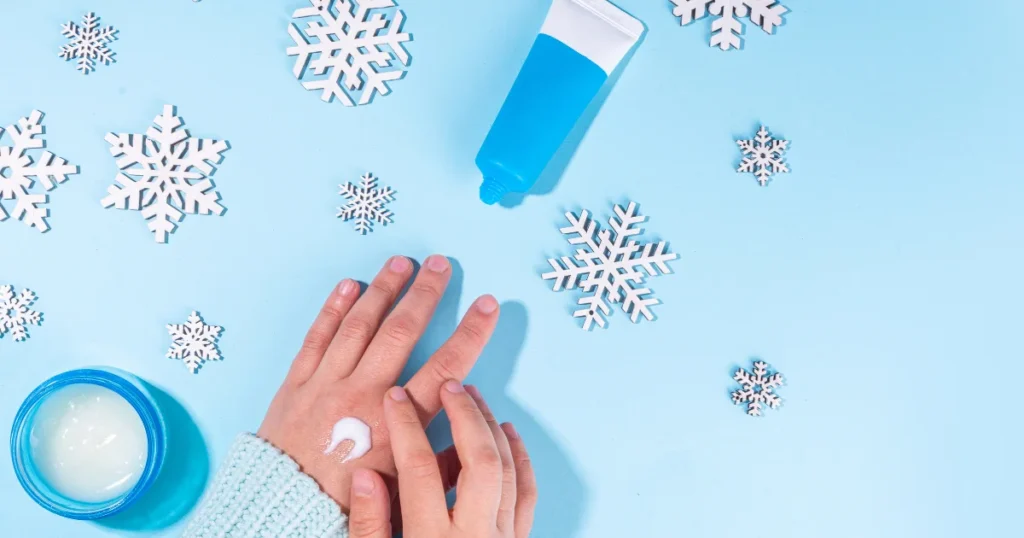 Persona echándose crema en las manos, sobre fondo azul y copos de nieve para destacar la importancia de cuidar la piel en invierno.